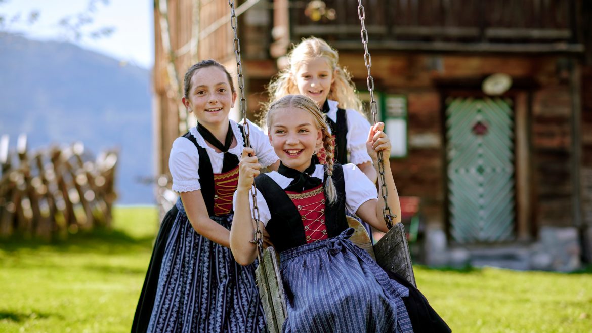 Drei lächelnde Mädchen in österreichischen Trachten, die an einem sonnigen Tag draußen in der Nähe eines rustikalen Holzhauses schaukeln und spielen.