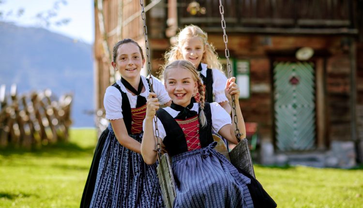 Drei lächelnde Mädchen in österreichischen Trachten, die an einem sonnigen Tag draußen in der Nähe eines rustikalen Holzhauses schaukeln und spielen.