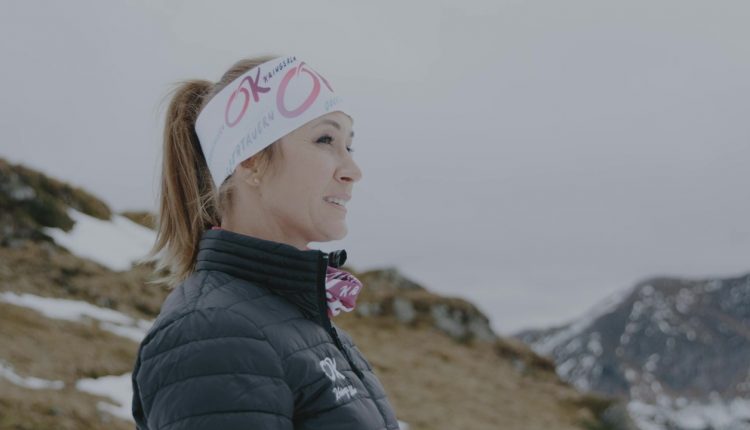Frau in Wintersportkleidung und Stirnband steht im Freien mit verschneiten Bergen im Hintergrund.