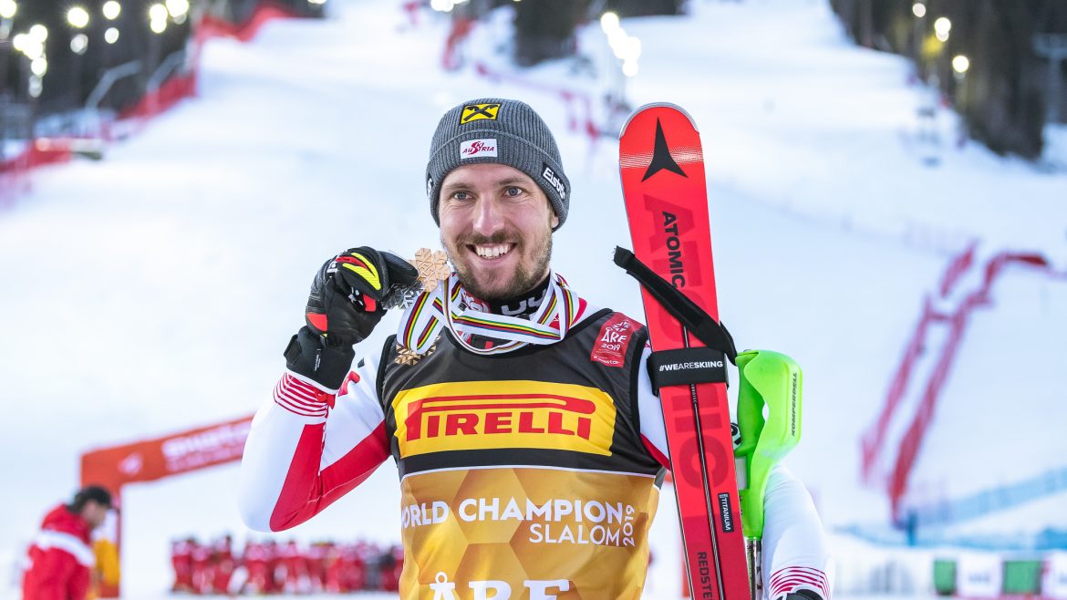 Ein Skifahrer in Rennkleidung lächelt, hält eine Medaille und Skier in der Hand, im Hintergrund eine verschneite Piste und Lichter.