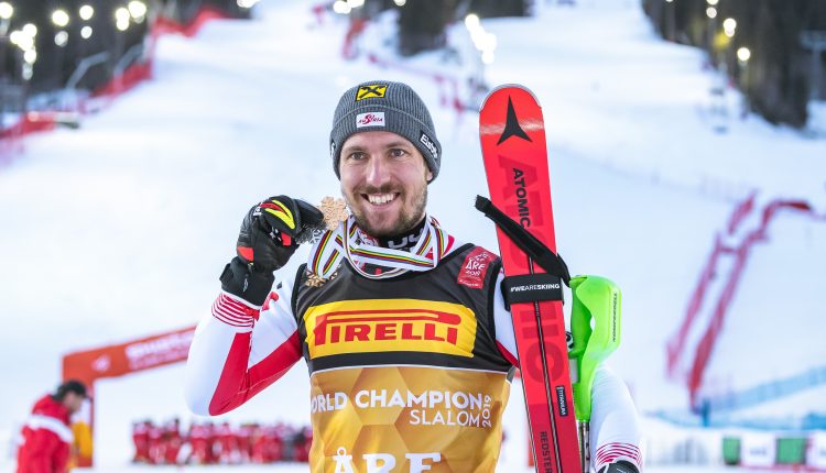 Ein Skifahrer in Rennkleidung lächelt, hält eine Medaille und Skier in der Hand, im Hintergrund eine verschneite Piste und Lichter.
