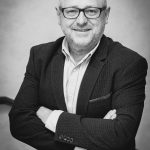 Ein lächelnder Mann mittleren Alters mit Brille und Anzugjacke steht mit verschränkten Armen.