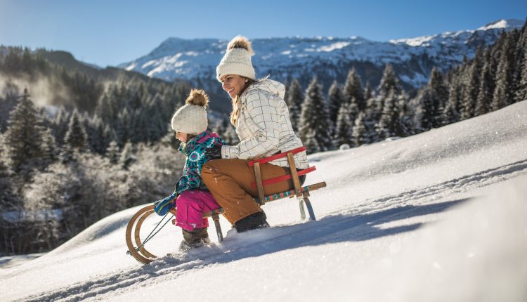 Frau und Kind rodeln einen verschneiten Hügel hinunter, mit Kiefern und Bergen im Hintergrund.