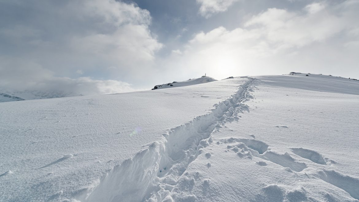 Ein schneebedeckter Weg mit Fußspuren führt unter einem wolkenverhangenen Himmel, durch den das Sonnenlicht bricht, einen sanften Hügel hinauf.