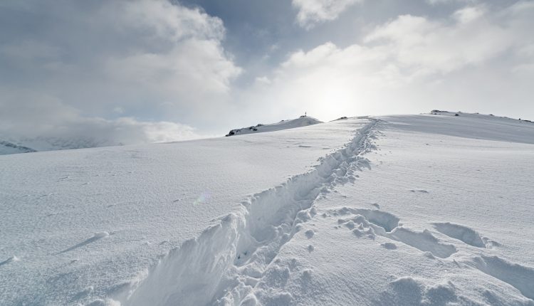 Ein schneebedeckter Weg mit Fußspuren führt unter einem wolkenverhangenen Himmel, durch den das Sonnenlicht bricht, einen sanften Hügel hinauf.