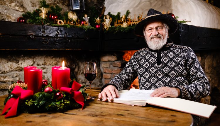 Ein lächelnder älterer Mann in einem festlichen Pullover liest an einem rustikalen Tisch mit Kerzen, Wein und Weihnachtsschmuck.