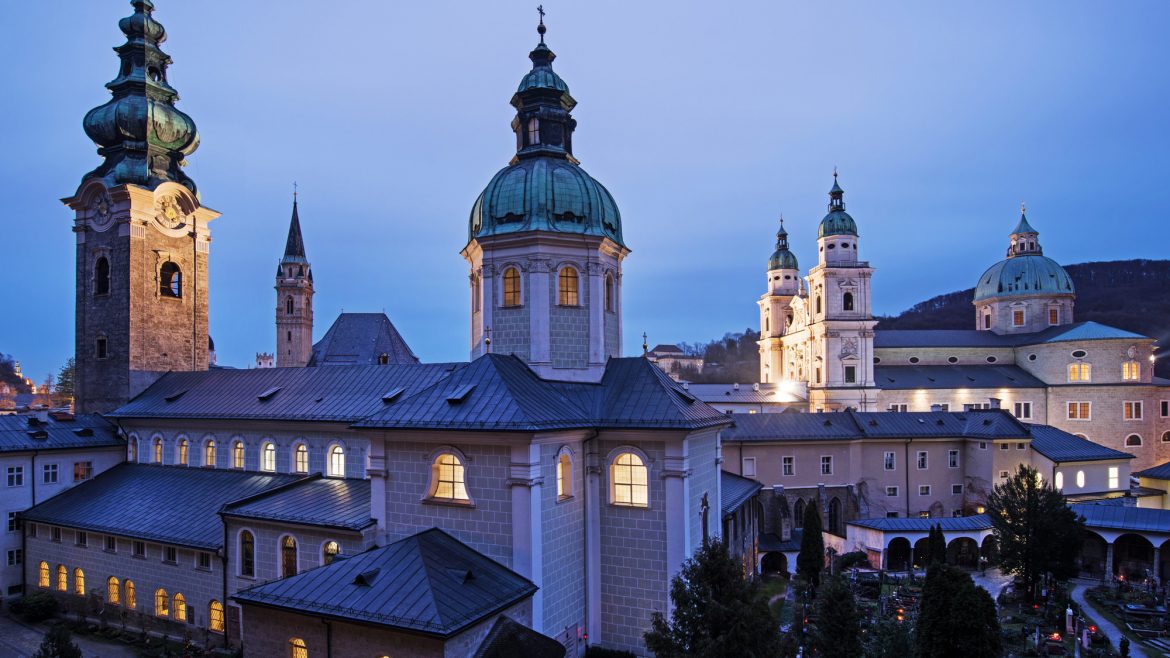 Historische Gebäude mit beleuchteten Kuppeln und Türmen in der Abenddämmerung in Salzburg, Österreich.