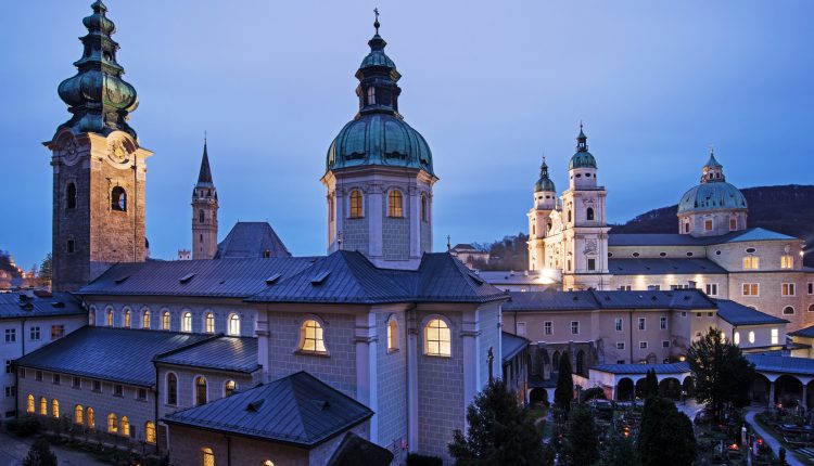 Historische Gebäude mit beleuchteten Kuppeln und Türmen in der Abenddämmerung in Salzburg, Österreich.