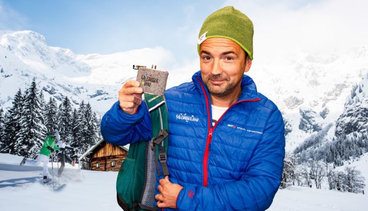 Ein Mann in blauer Jacke und grüner Mütze hält eine Snackbar in den verschneiten Bergen mit einer Hütte und Skifahrern im Hintergrund.