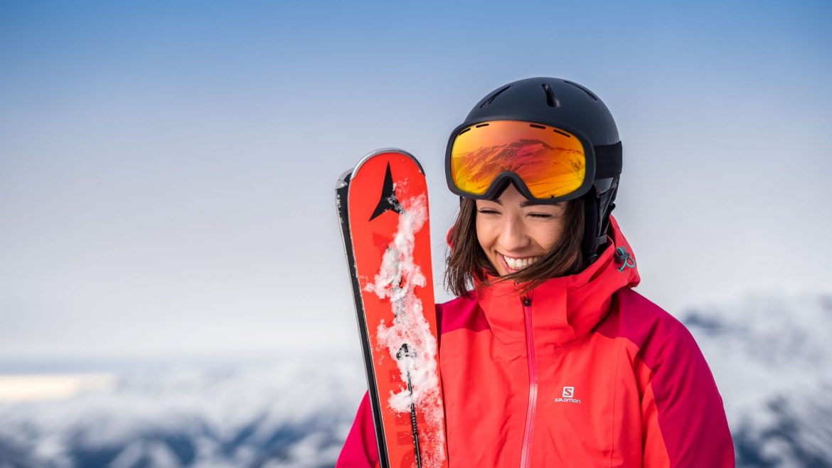 Frau in roter Skijacke und Helm, lächelnd, mit Skiern in der Hand, mit verschneiten Bergen im Hintergrund.