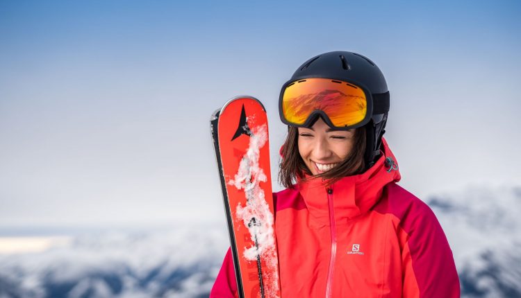 Frau in roter Skijacke und Helm, lächelnd, mit Skiern in der Hand, mit verschneiten Bergen im Hintergrund.
