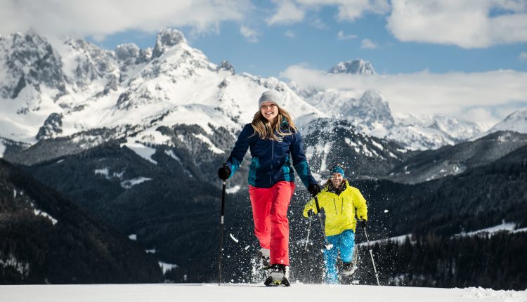 Zwei Personen beim Schneeschuhwandern auf einem verschneiten Berg mit malerischen Gipfeln und Wäldern im Hintergrund.