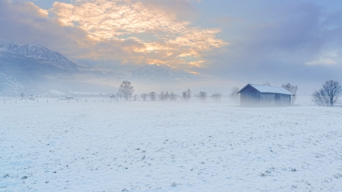 Ein schneebedecktes Feld mit einer kleinen Scheune, Bäumen und Bergen unter einem wolkenverhangenen Himmel bei Sonnenaufgang.