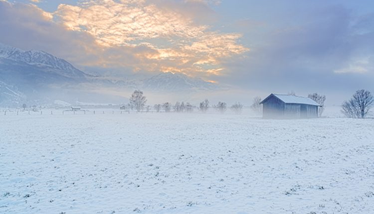 Ein schneebedecktes Feld mit einer kleinen Scheune, Bäumen und Bergen unter einem wolkenverhangenen Himmel bei Sonnenaufgang.