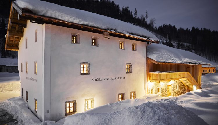 Ein schneebedecktes Museumsgebäude leuchtet nachts warm in einer verschneiten, bewaldeten Berglandschaft.