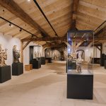 Geräumige Museumsgalerie mit Holzbalken, in der Skulpturen in Vitrinen auf hellem Holzboden ausgestellt sind.