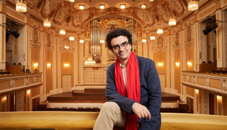 Ein Mann mit blauem Blazer und rotem Schal sitzt in einem prunkvollen Konzertsaal mit Kronleuchtern und einer großen Pfeifenorgel.