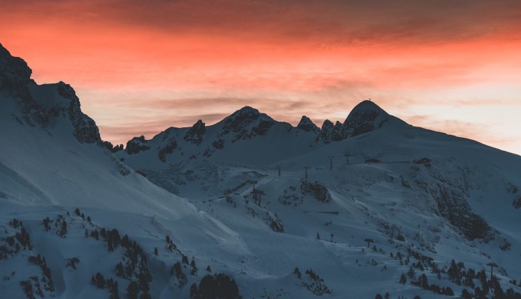 Verschneite Berggipfel mit Skiliften bei Sonnenuntergang unter einem dramatischen orange-grauen Himmel.