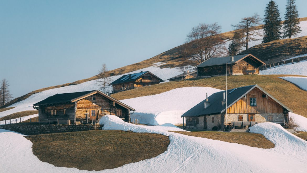 Holzhütten an einem verschneiten Hang mit Grasflächen und ein paar Bäumen unter einem klaren Himmel.