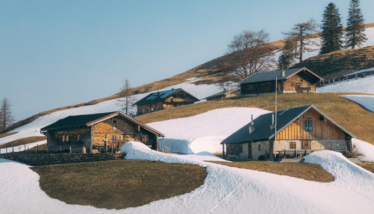 Holzhütten an einem verschneiten Hang mit Grasflächen und ein paar Bäumen unter einem klaren Himmel.