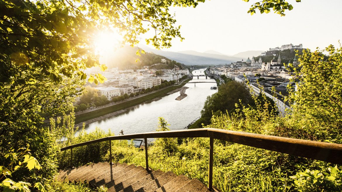Eine geschwungene Steintreppe führt hinunter ins Grüne, mit Blick auf eine sonnenbeschienene Stadt am Fluss und die fernen Berge.