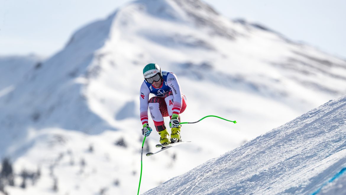 Ein Skifahrer in der Luft rast auf einem verschneiten Berg mit sichtbaren Gipfeln im Hintergrund bergab.