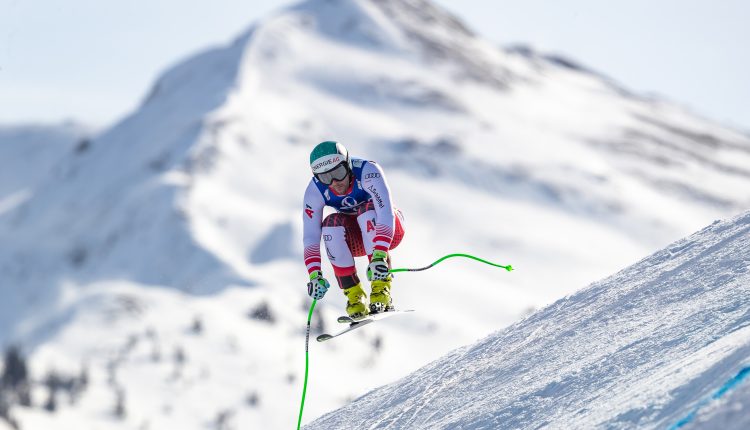 Ein Skifahrer in der Luft rast auf einem verschneiten Berg mit sichtbaren Gipfeln im Hintergrund bergab.