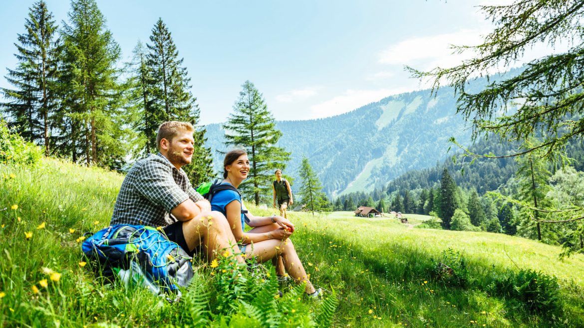 Zwei Personen ruhen sich lächelnd auf einem grasbewachsenen Berghang aus, mit Bäumen und fernen Bergen im Hintergrund.
