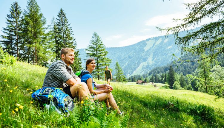 Zwei Personen ruhen sich lächelnd auf einem grasbewachsenen Berghang aus, mit Bäumen und fernen Bergen im Hintergrund.