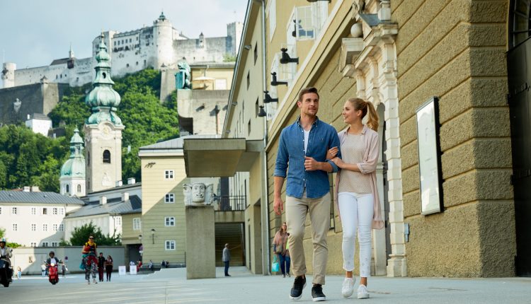 Ein Paar spaziert gemeinsam auf einer Straße in der Stadt mit historischen Gebäuden und einer Festung im Hintergrund.