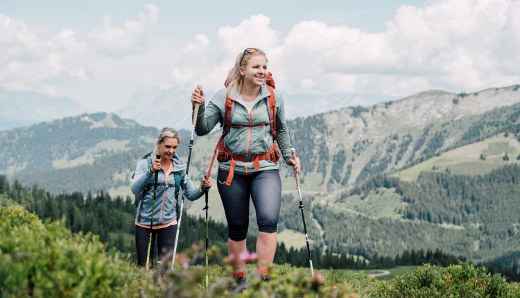 Zwei Frauen wandern mit Trekkingstöcken auf einem grünen Bergpfad mit malerischer Aussicht im Hintergrund.