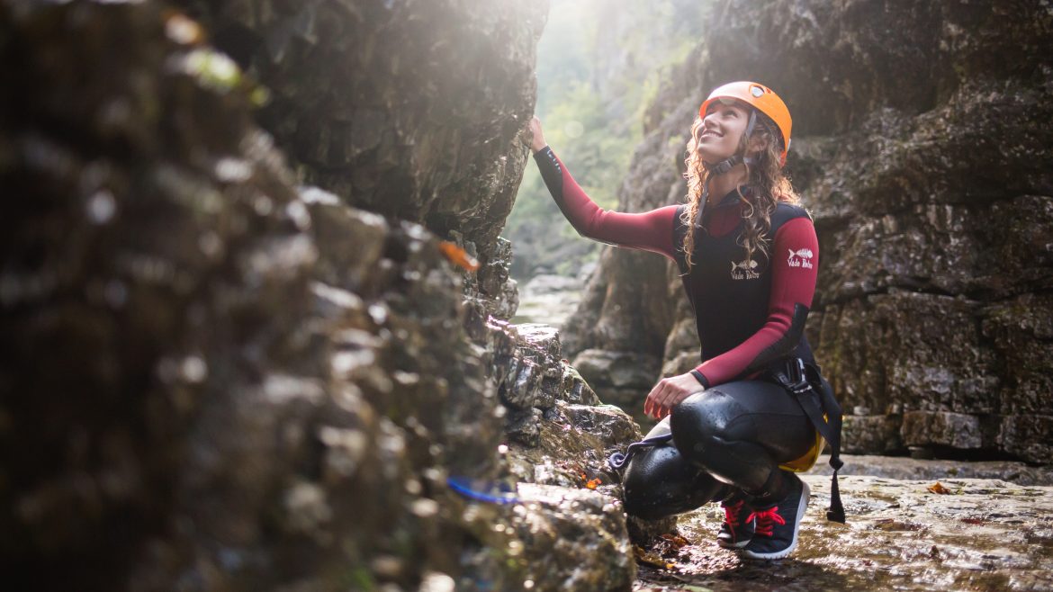 Eine Frau in Outdoor-Ausrüstung und mit Helm kniet auf felsigem Boden und berührt einen Felsblock in einer sonnenbeschienenen Schlucht.