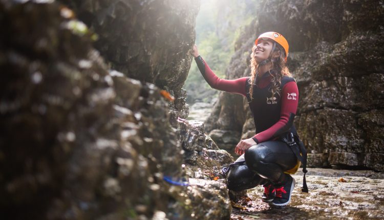 Eine Frau in Outdoor-Ausrüstung und mit Helm kniet auf felsigem Boden und berührt einen Felsblock in einer sonnenbeschienenen Schlucht.