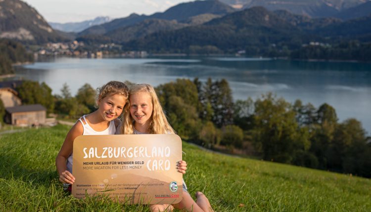 Zwei lächelnde Mädchen sitzen im Gras an einem See und halten ein Schild, das für das SalzburgerLand in Österreich wirbt.