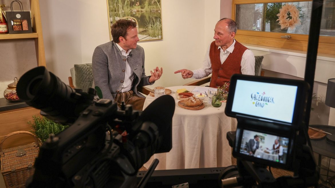 Zwei Männer in traditioneller Kleidung unterhalten sich an einem kleinen Tisch, gefilmt von einer Kamera in einem gemütlichen Raum.