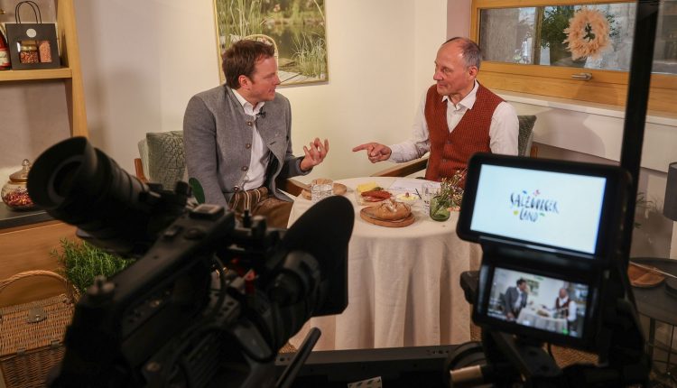 Zwei Männer in traditioneller Kleidung unterhalten sich an einem kleinen Tisch, gefilmt von einer Kamera in einem gemütlichen Raum.