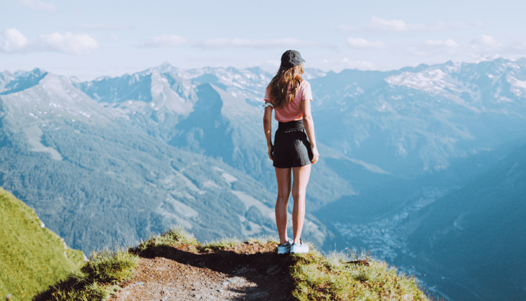 Eine Frau steht auf einer grasbewachsenen Klippe mit Blick auf die fernen Berge unter einem klaren blauen Himmel.