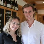 Ein lächelnder Mann und eine lächelnde Frau stehen zusammen in einem Haus in der Nähe von Kreidetafel-Menüs und Weinflaschen.