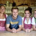 Vier lächelnde Erwachsene, drei Frauen in traditioneller österreichischer Kleidung und ein Mann, der sich auf einen Holztresen stützt.