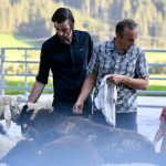 Zwei Männer und ein Junge stehen zwischen Schafen im Freien, mit Bäumen und einem Zaun im Hintergrund.