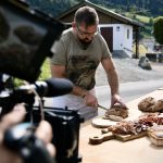 Ein Mann schneidet an einem Tisch mit Fleisch und Brot im Freien Brot auf, während eine Kamera ihn in einer ländlichen Umgebung filmt.