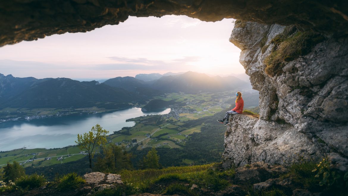 Eine Person sitzt auf einem Felsvorsprung mit Blick auf ein malerisches Tal, einen See und Berge bei Sonnenuntergang.