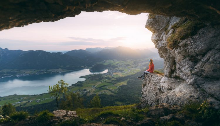 Eine Person sitzt auf einem Felsvorsprung mit Blick auf ein malerisches Tal, einen See und Berge bei Sonnenuntergang.