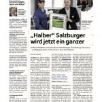 Zwei Männer stehen lächelnd da und schütteln sich die Hände, während sie eine gerahmte Urkunde bei einer Indoor-Veranstaltung halten.