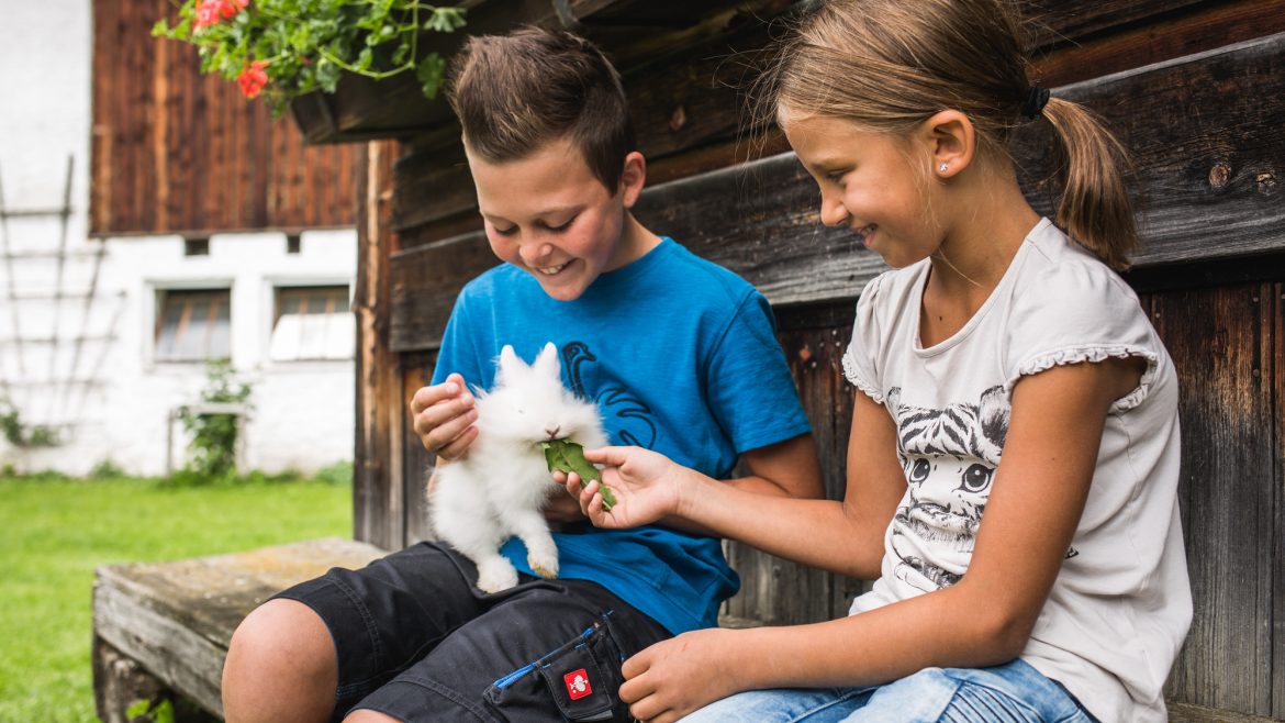 Zwei Kinder sitzen auf einer Bank, lächeln und füttern ein weißes Kaninchen mit einem grünen Blatt vor einem Holzhaus.