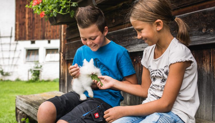 Zwei Kinder sitzen auf einer Bank, lächeln und füttern ein weißes Kaninchen mit einem grünen Blatt vor einem Holzhaus.
