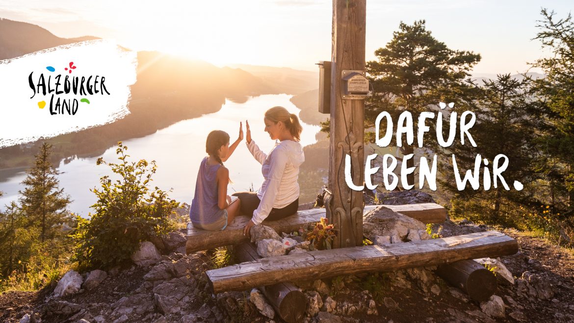 Eine Frau und ein Kind geben sich bei Sonnenuntergang an einem Holzkreuz auf einem Berg mit Blick auf einen See die Hand.