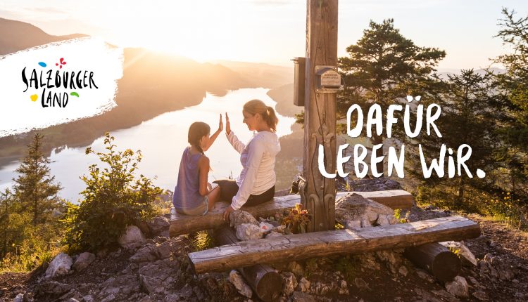 Eine Frau und ein Kind geben sich bei Sonnenuntergang an einem Holzkreuz auf einem Berg mit Blick auf einen See die Hand.