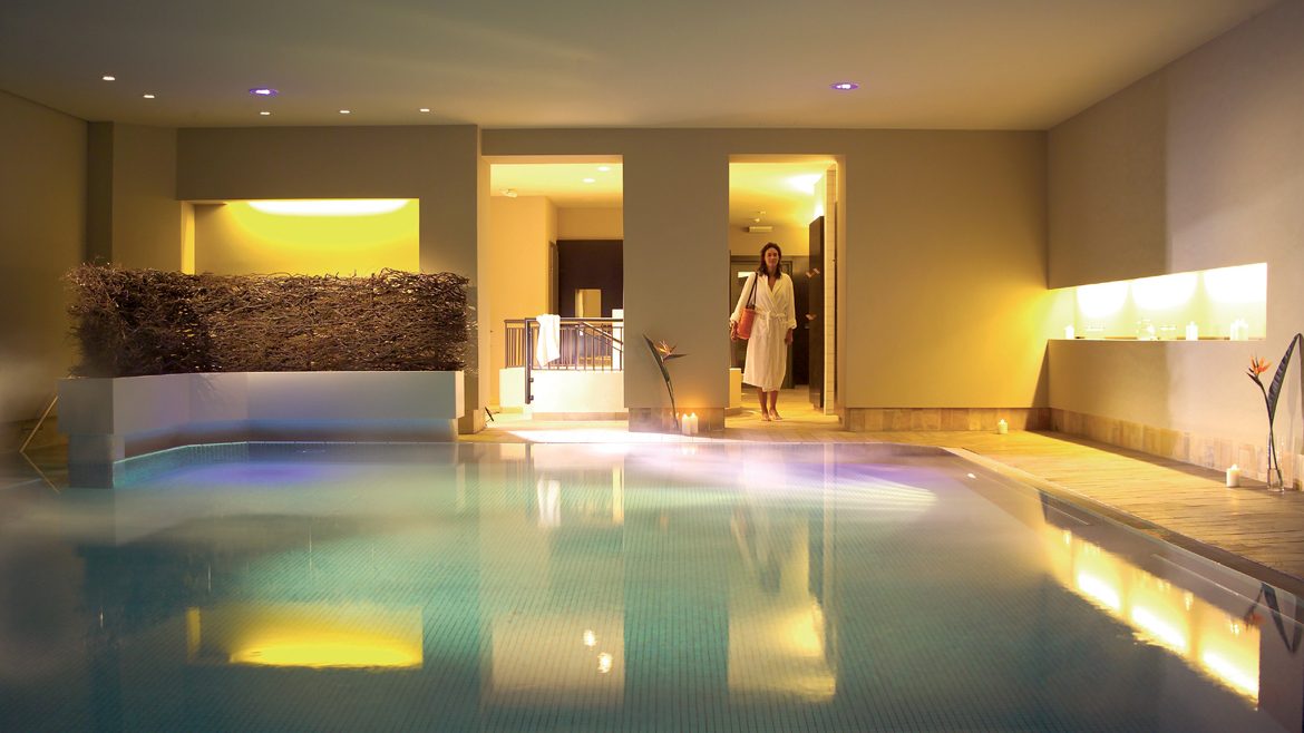 Eine Frau in einem weißen Bademantel betritt einen Indoor-Spa-Pool mit warmer Beleuchtung und modernem Dekor.