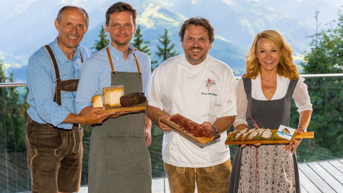Vier lächelnde Menschen in traditioneller österreichischer Alpentracht halten Teller mit Essen, während im Hintergrund die Berge zu sehen sind.